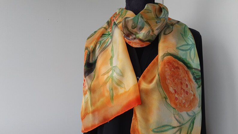 May include: A silk scarf with a hand-painted design of orange and green fruits and leaves. The scarf is a light orange color with a gradient of orange and red at the bottom.