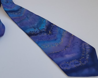 Pode incluir: Uma gravata de seda azul com um padrão de redemoinho de ouro e roxo. A gravata tem um efeito aquarela sutil.