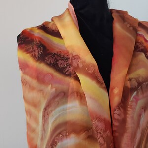 May include: A long, flowing silk scarf with a hand-painted design in shades of orange, yellow, brown, and red. The scarf features a swirling, abstract pattern that resembles clouds or waves.