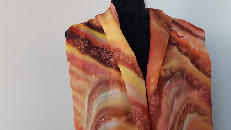 May include: A long, flowing silk scarf with a hand-painted design in shades of orange, yellow, brown, and red. The scarf features a swirling, abstract pattern that resembles clouds or waves.