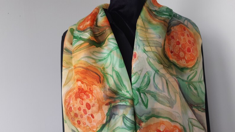 May include: A silk scarf with a hand-painted design of orange and green fruit with leaves. The scarf is a light beige color with a watercolor effect.