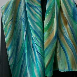 May include: A silk scarf with a hand-painted design of blue, green, and gold stripes. The scarf is folded in half and draped over a black surface.