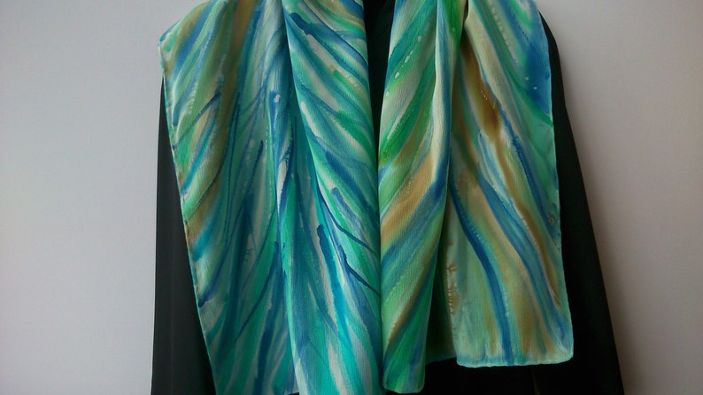 May include: A silk scarf with a hand-painted design of blue, green, and gold stripes. The scarf is folded in half and draped over a black surface.