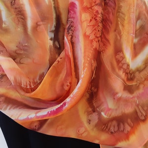 May include: A silk scarf with a hand-painted design in shades of orange, brown, and pink. The scarf is folded and draped over a black surface.