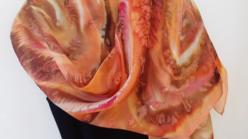 May include: A silk scarf with a hand-painted design in shades of orange, brown, and pink. The scarf is folded and draped over a black surface.