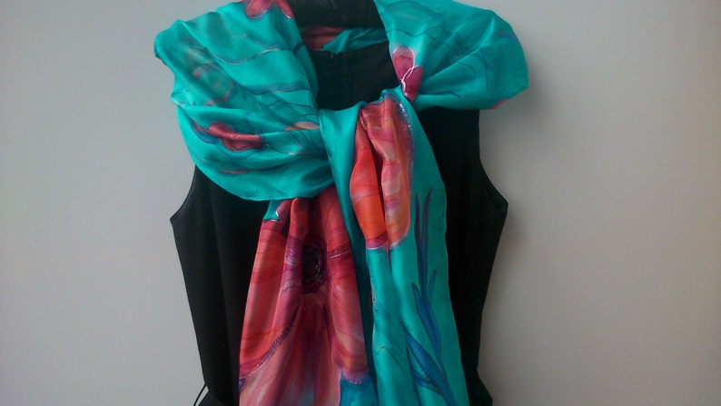 May include: A turquoise silk scarf with a floral design in shades of pink, orange, and red. The scarf is tied around the neck of a black sleeveless dress.