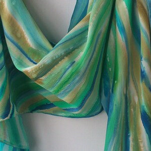 May include: A green, blue, and white silk scarf with a hand-painted abstract design. The scarf is made of soft, flowing fabric and has a delicate, ethereal feel.