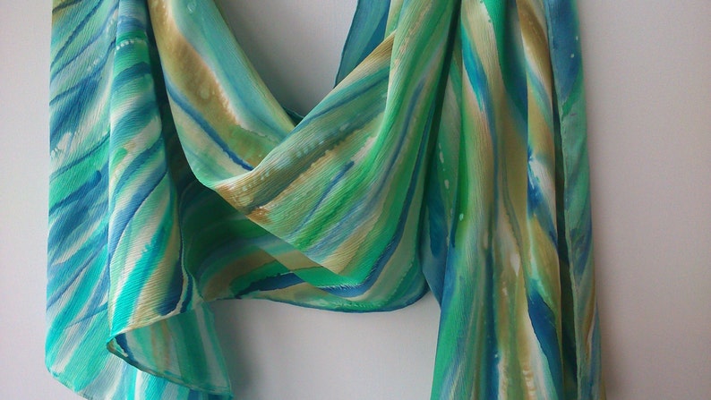 May include: A green, blue, and white silk scarf with a hand-painted abstract design. The scarf is made of soft, flowing fabric and has a delicate, ethereal feel.