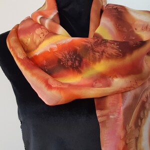 May include: A long, flowing silk scarf with a hand-painted design in shades of orange, yellow, and brown. The scarf is draped around a mannequin's neck.