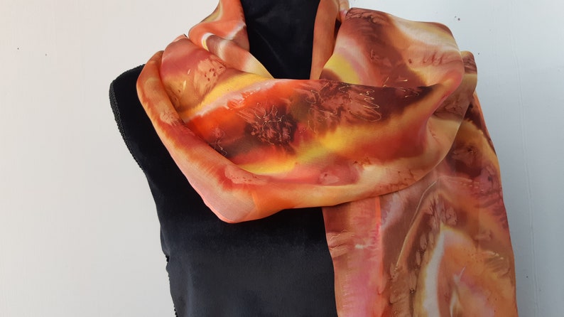 May include: A long, flowing silk scarf with a hand-painted design in shades of orange, yellow, and brown. The scarf is draped around a mannequin's neck.