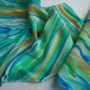 May include: A silk scarf with a hand-painted design of blue, green, and gold stripes. The scarf is folded and draped on a white surface.