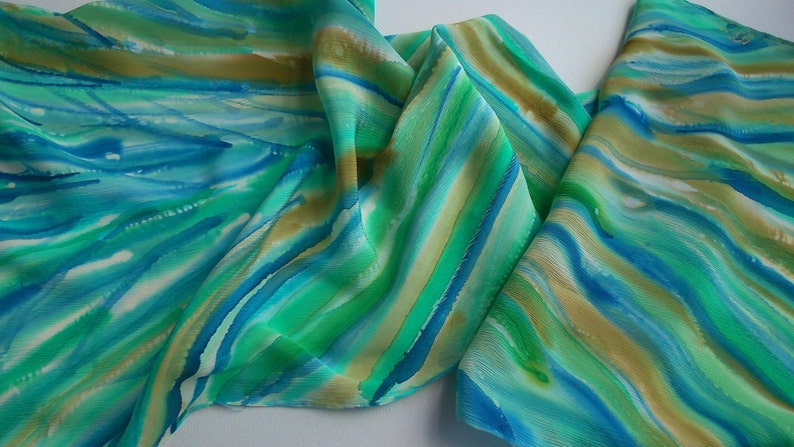 May include: A silk scarf with a hand-painted design of blue, green, and gold stripes. The scarf is folded and draped on a white surface.