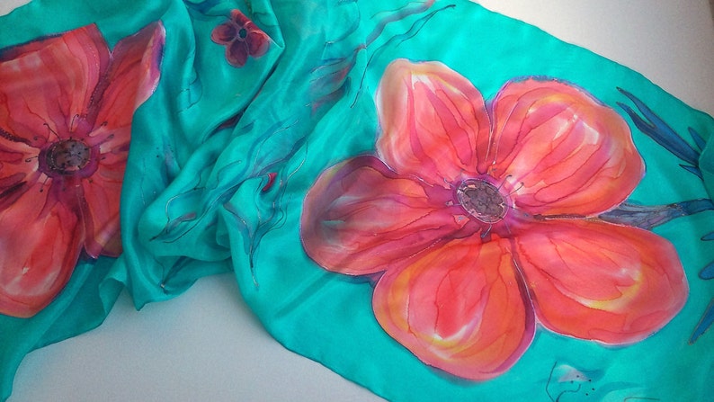 May include: A turquoise silk scarf with hand-painted red and orange flowers. The scarf features a floral design with delicate details.