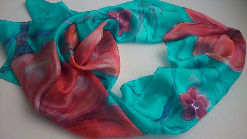 May include: A turquoise silk scarf with a hand-painted design of red and pink flowers. The scarf is folded in half and the design is visible on both sides.