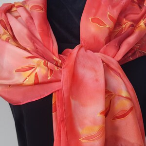 May include: A red silk scarf with a hand-painted design of orange and yellow leaves. The scarf is tied around a person's neck.