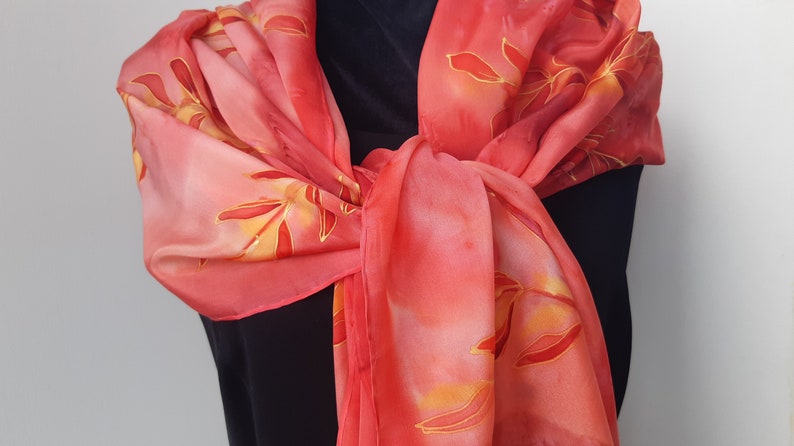 May include: A red silk scarf with a hand-painted design of orange and yellow leaves. The scarf is tied around a person's neck.
