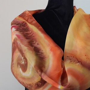 May include: A silk scarf with a swirling pattern in shades of orange, yellow, and brown. The scarf is draped over a black mannequin.