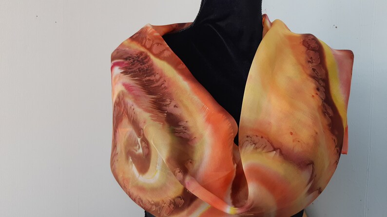 May include: A silk scarf with a swirling pattern in shades of orange, yellow, and brown. The scarf is draped over a black mannequin.