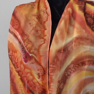 May include: A long, flowing silk scarf with a hand-painted design in shades of orange, brown, and yellow. The design features swirling patterns and abstract shapes.
