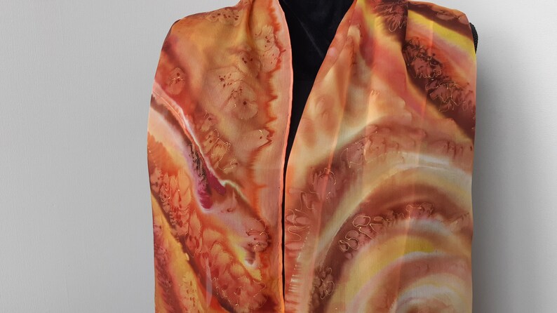 May include: A long, flowing silk scarf with a hand-painted design in shades of orange, brown, and yellow. The design features swirling patterns and abstract shapes.