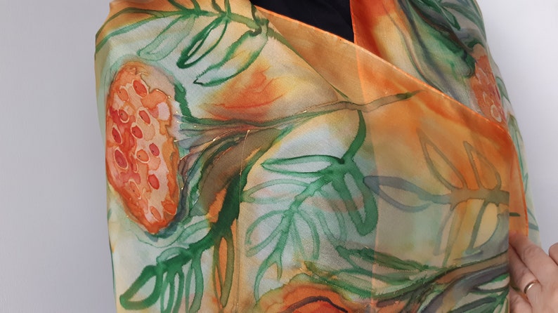 May include: A silk scarf with a watercolor design of orange and green flowers and leaves. The scarf is folded and draped over a person's shoulder.