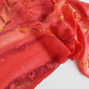 May include: A red silk scarf with a hand-painted design of golden branches and leaves. The scarf has a gradient of color from a darker red to a lighter orange. The artist's signature is visible in the bottom left corner.