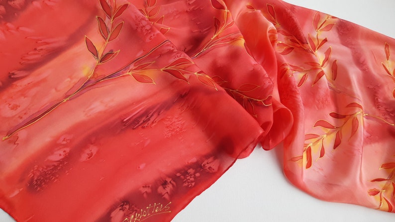 May include: A red silk scarf with a hand-painted design of golden branches and leaves. The scarf has a gradient of color from a darker red to a lighter orange. The artist's signature is visible in the bottom left corner.