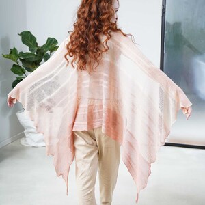 May include: A light pink, open-front shawl with a flowing, asymmetrical design. The shawl features a gradient effect, transitioning from a pale pink to a deeper coral hue. The model is wearing beige pants and shoes.