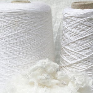 Puede incluir: Dos carretes de hilo blanco, uno con una textura lisa y el otro con una textura m&aacute;s texturizada, similar a una cuerda. Un mont&oacute;n de fibra de algod&oacute;n blanco se encuentra en primer plano.