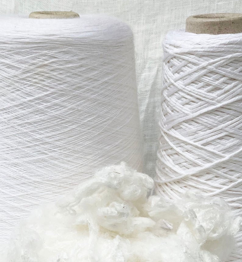 Puede incluir: Dos carretes de hilo blanco, uno con una textura lisa y el otro con una textura m&aacute;s texturizada, similar a una cuerda. Un mont&oacute;n de fibra de algod&oacute;n blanco se encuentra en primer plano.