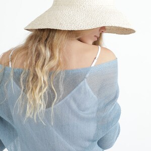 Op de afbeelding: Een strohoed met brede rand in lichtbeige. De hoed wordt gedragen met een lichtblauwe, doorschijnende, off-the-shoulder top. Het model heeft lang, golvend blond haar. De achtergrond is wit.