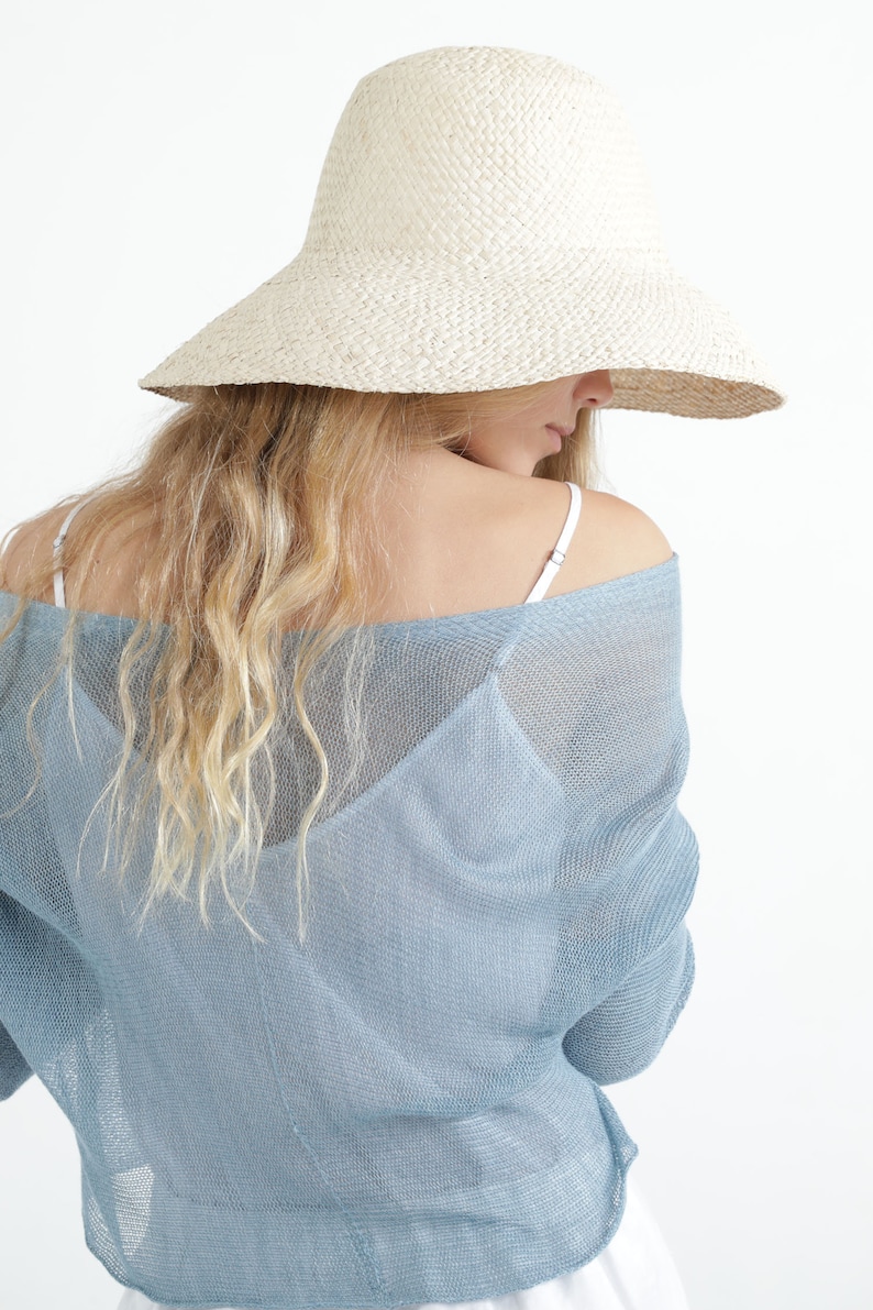 Op de afbeelding: Een strohoed met brede rand in lichtbeige. De hoed wordt gedragen met een lichtblauwe, doorschijnende, off-the-shoulder top. Het model heeft lang, golvend blond haar. De achtergrond is wit.