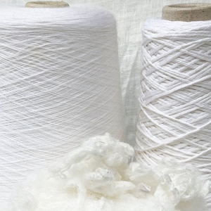Puede incluir: Dos carretes de hilo blanco, uno con una textura lisa y el otro con una textura m&aacute;s texturizada, similar a una cuerda. Un mont&oacute;n de fibra de algod&oacute;n blanco se encuentra en primer plano.