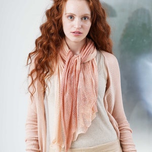 May include: A woman wearing a light peach-colored scarf and cardigan. The scarf is draped around the neck and has a soft, flowing appearance. The cardigan is a matching color and complements the scarf. The woman is also wearing a light-colored top and pants.