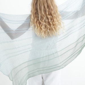 May include: A sheer, light blue and green striped shawl. The shawl is held open, displaying its delicate weave and color gradient. The fabric appears lightweight and airy, suitable for layering or as a decorative accessory.