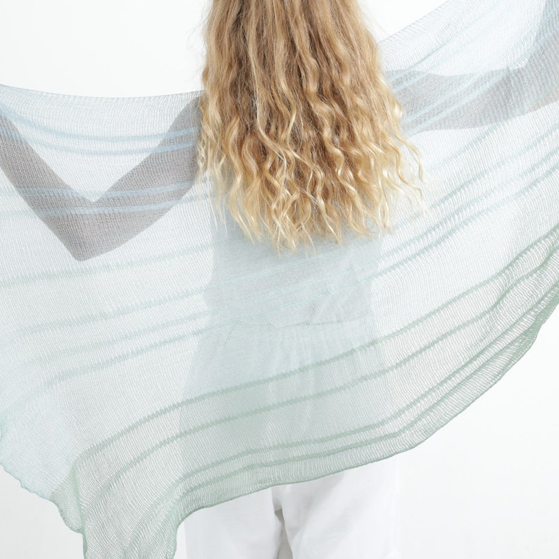 May include: A sheer, light blue and green striped shawl. The shawl is held open, displaying its delicate weave and color gradient. The fabric appears lightweight and airy, suitable for layering or as a decorative accessory.