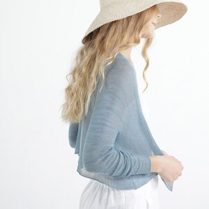 Op de afbeelding: Een lichtblauwe bolero-stijl vest met open voorkant en lange mouwen. Het model draagt een breedgerande strohoed en een witte broek. Het vest is gemaakt van een lichte, transparante stof, perfect om te dragen.