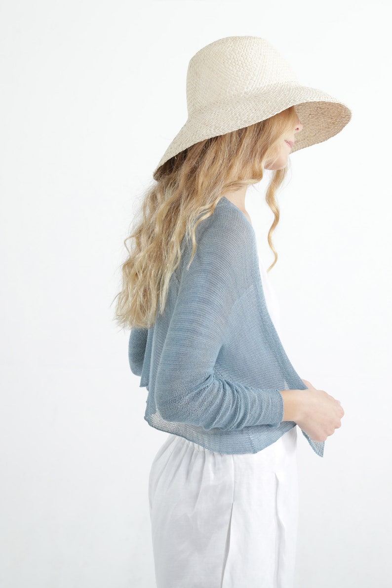Op de afbeelding: Een lichtblauwe bolero-stijl vest met open voorkant en lange mouwen. Het model draagt een breedgerande strohoed en een witte broek. Het vest is gemaakt van een lichte, transparante stof, perfect om te dragen.