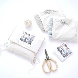 Op de afbeelding: Een bovenaanzicht van een verzameling items: een witte kussensloop met een doorzichtige hoes, twee stoffen zakken, een bol touw, een schaar en visitekaartjes. De kussensloop heeft een collage van afbeeldingen en de tekst "TOOSHAYA".