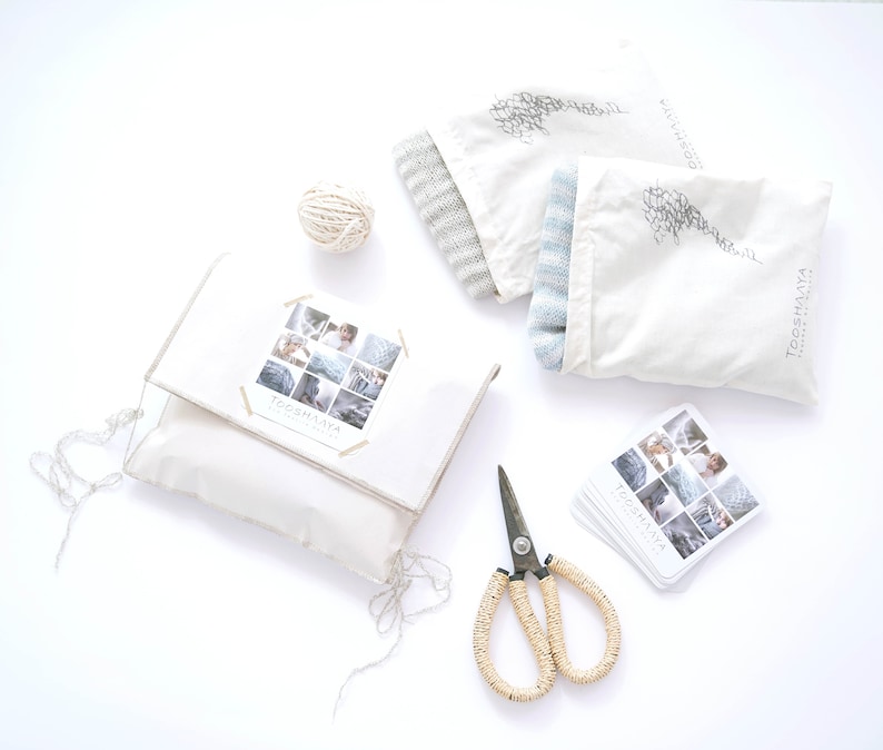Op de afbeelding: Een bovenaanzicht van een verzameling items: een witte kussensloop met een doorzichtige hoes, twee stoffen zakken, een bol touw, een schaar en visitekaartjes. De kussensloop heeft een collage van afbeeldingen en de tekst "TOOSHAYA".
