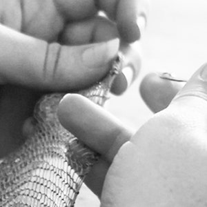 May include: Close-up, black and white image of a person's hands manipulating a small, intricately designed object. The object appears to be made of a mesh-like material, possibly metal or a similar substance. The hands are in focus, highlighting the detail of the object.