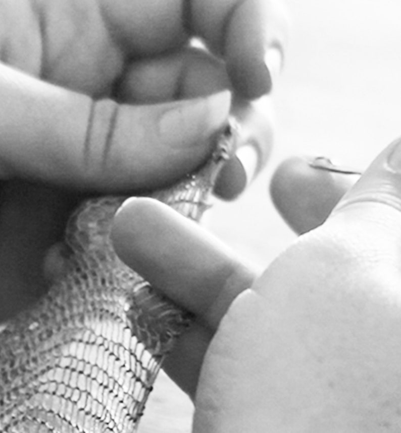 May include: Close-up, black and white image of a person's hands manipulating a small, intricately designed object. The object appears to be made of a mesh-like material, possibly metal or a similar substance. The hands are in focus, highlighting the detail of the object.