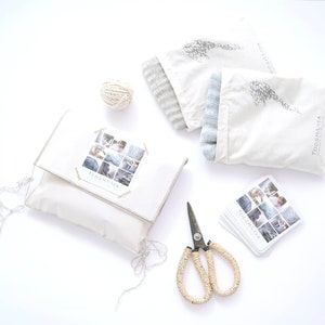 Puede incluir: Una bolsa de tela blanca con un gorro de punto gris y azul en su interior. La bolsa tiene el texto "TOOSHANA" impreso. Hay una bola de hilo, unas tijeras y una tarjeta con fotos de art&iacute;culos de punto sobre una superficie blanca.