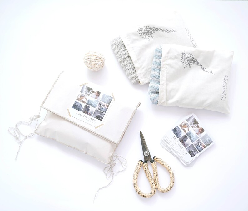 Puede incluir: Una bolsa de tela blanca con un gorro de punto gris y azul en su interior. La bolsa tiene el texto "TOOSHANA" impreso. Hay una bola de hilo, unas tijeras y una tarjeta con fotos de art&iacute;culos de punto sobre una superficie blanca.