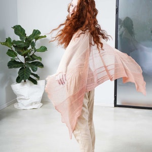 May include: A light peach-colored shawl with a flowing, open design. The shawl is draped over a person wearing beige pants and shoes. The person has long, red hair. A large potted plant is in the background.