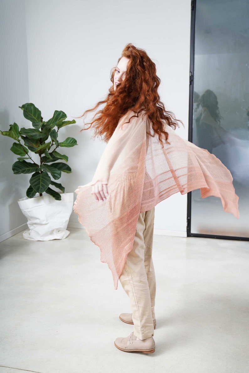May include: A light peach-colored shawl with a flowing, open design. The shawl is draped over a person wearing beige pants and shoes. The person has long, red hair. A large potted plant is in the background.