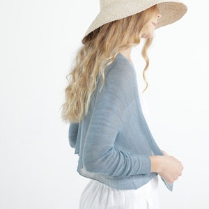 Op de afbeelding: Een vrouw draagt een lichtblauwe bolero-cardigan met lange mouwen en een breedgerande strohoed. Ze draagt ook een witte broek. De outfit staat tegen een witte achtergrond.