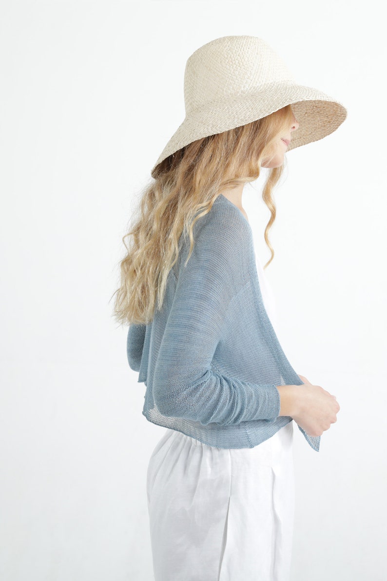 Op de afbeelding: Een vrouw draagt een lichtblauwe bolero-cardigan met lange mouwen en een breedgerande strohoed. Ze draagt ook een witte broek. De outfit staat tegen een witte achtergrond.