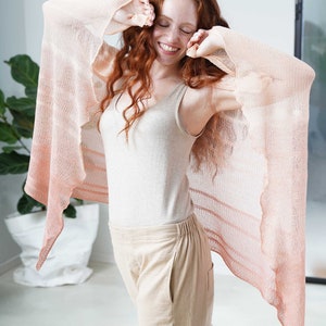 May include: A light peach-colored shawl with a flowing design. The shawl is draped over the shoulders and arms. The person is wearing a beige tank top and beige pants.