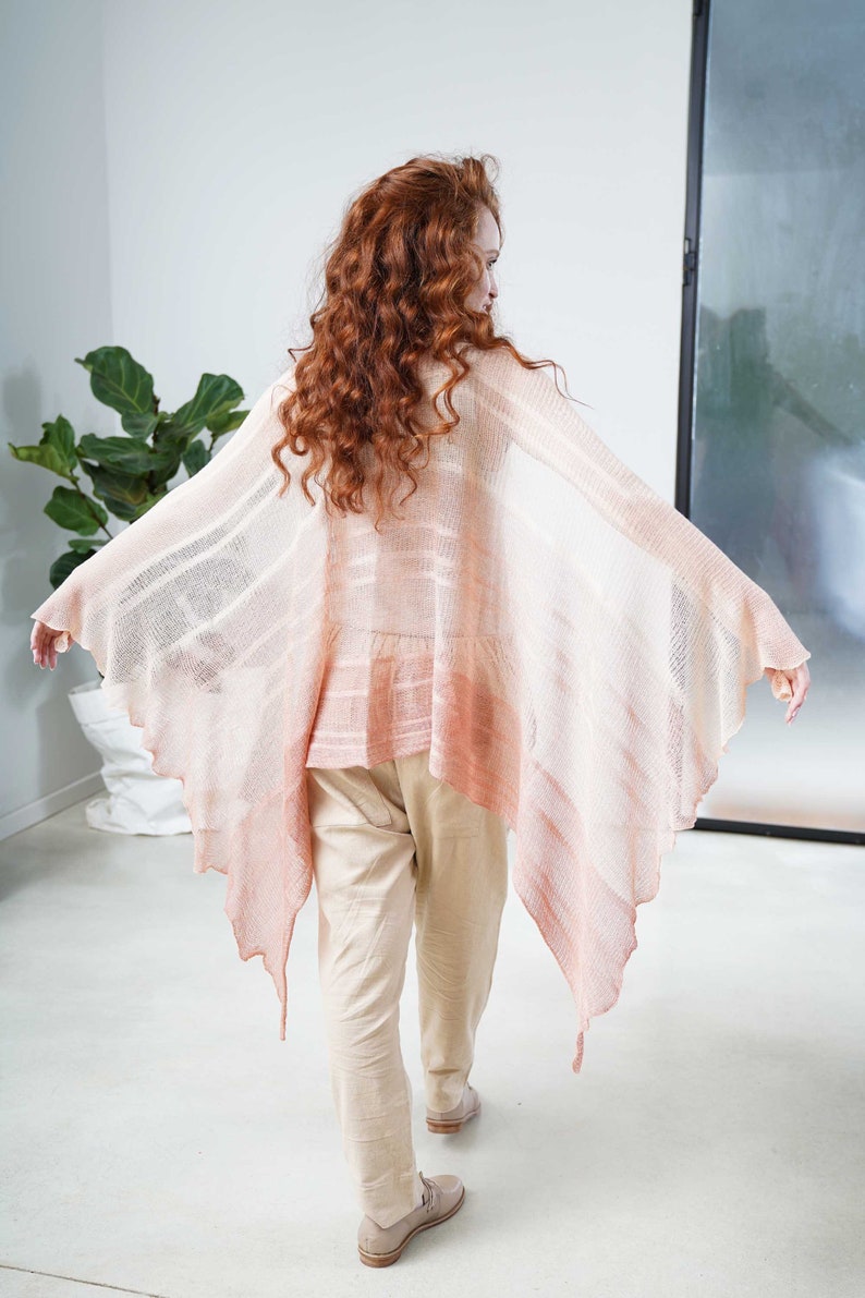 May include: A light pink, open-front shawl with a flowing, asymmetrical design. The shawl features a gradient effect, transitioning from a pale pink to a deeper coral hue. The model is wearing beige pants and shoes.